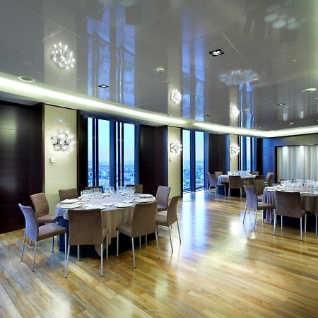 Hotel Eurostars Madrid Tower Esterno foto The photo depicts a modern dining room or banquet hall with large windows that provide a view of the outside. The room features elegant wooden flooring and contemporary furniture, including round tables set with plates, glasses, and cutlery. Various 