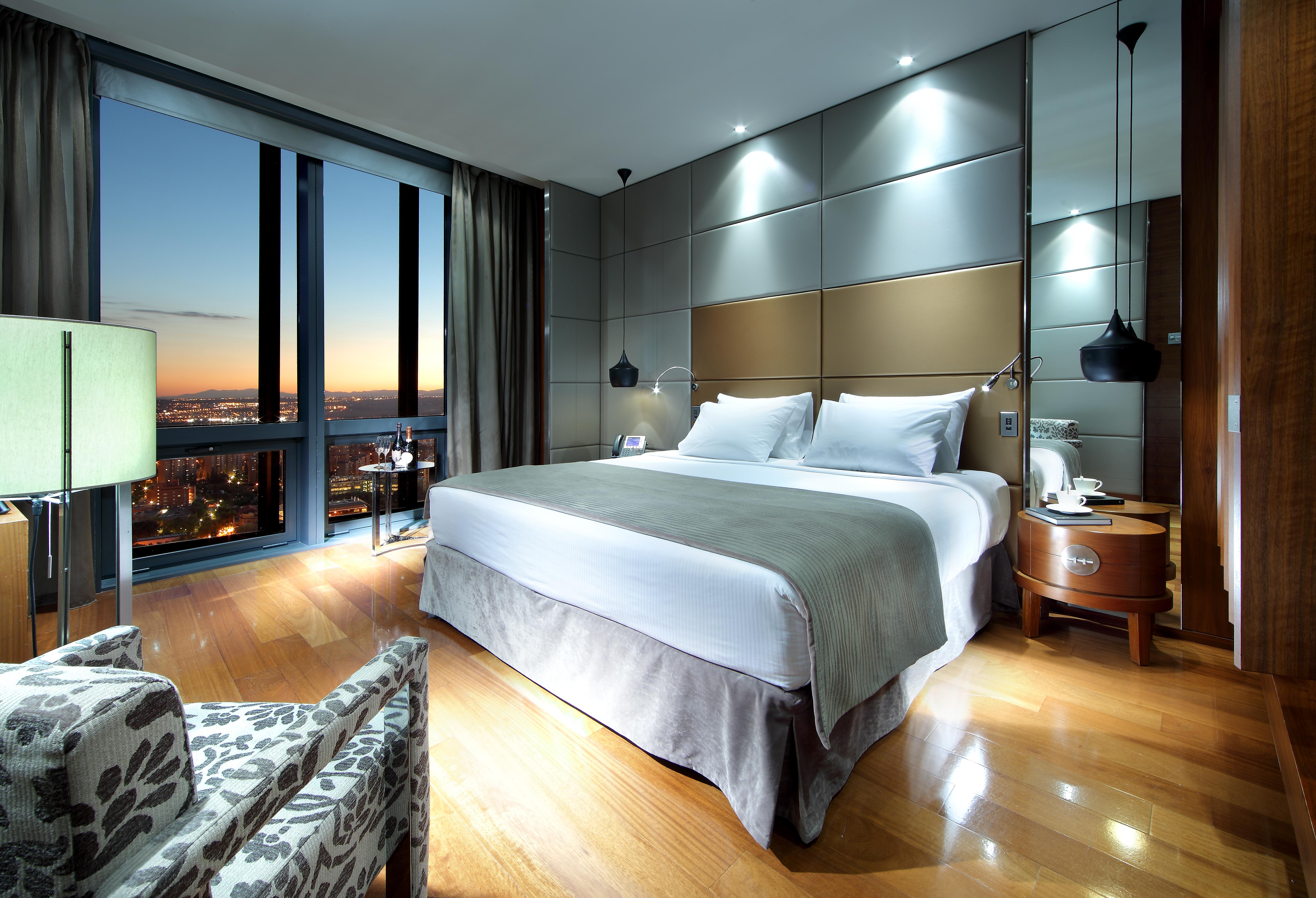 Hotel Eurostars Madrid Tower Esterno foto The image shows a modern hotel room featuring a large bed with white linens and a gray throw. There are stylish bedside lamps and a sleek nightstand. A comfortable chair with a patterned fabric sits to the side. The room has large windows that reveal