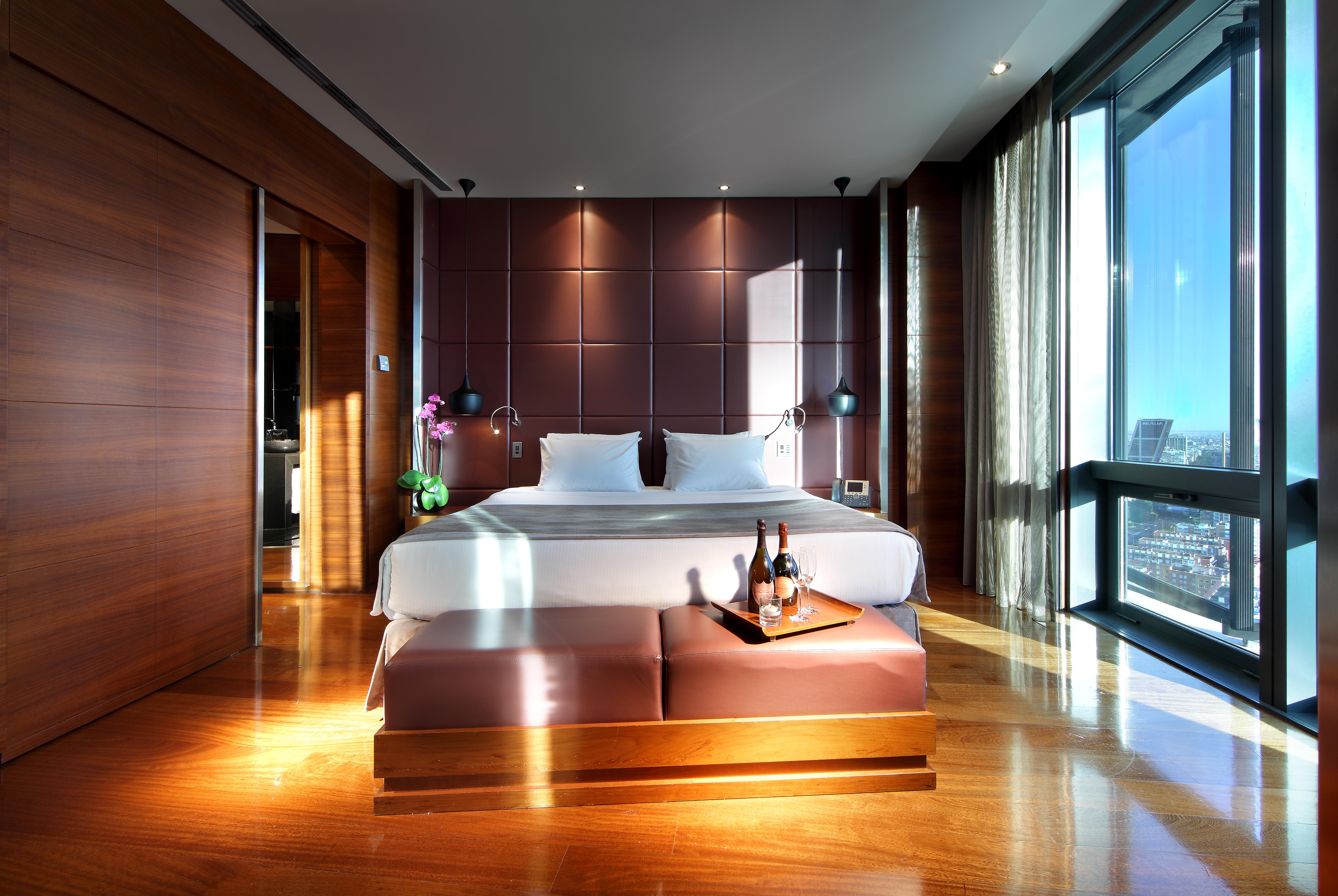 Hotel Eurostars Madrid Tower Esterno foto The photo shows a modern hotel room featuring a large bed with white linens positioned against a stylish dark brown accent wall. There are two bedside lamps on either side of the bed, and a small table at the foot of the bed holding a bottle of wine 