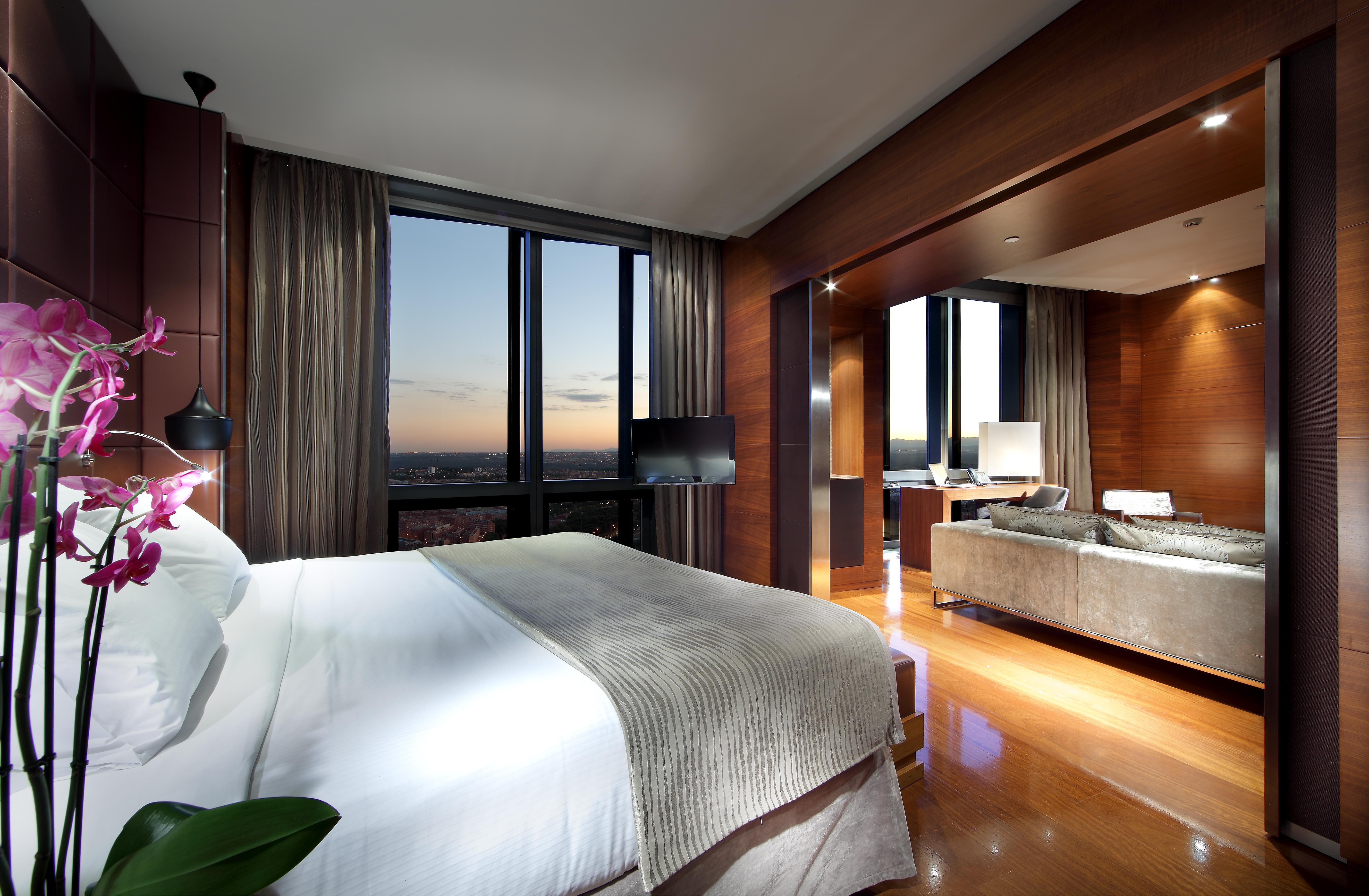 Hotel Eurostars Madrid Tower Esterno foto The photo depicts a modern hotel room interior. It features a spacious layout with a large bed dressed in white linens, accompanied by a subtle patterned blanket. To the side, there is a small decorative plant. 

The room is illuminated by ambient li