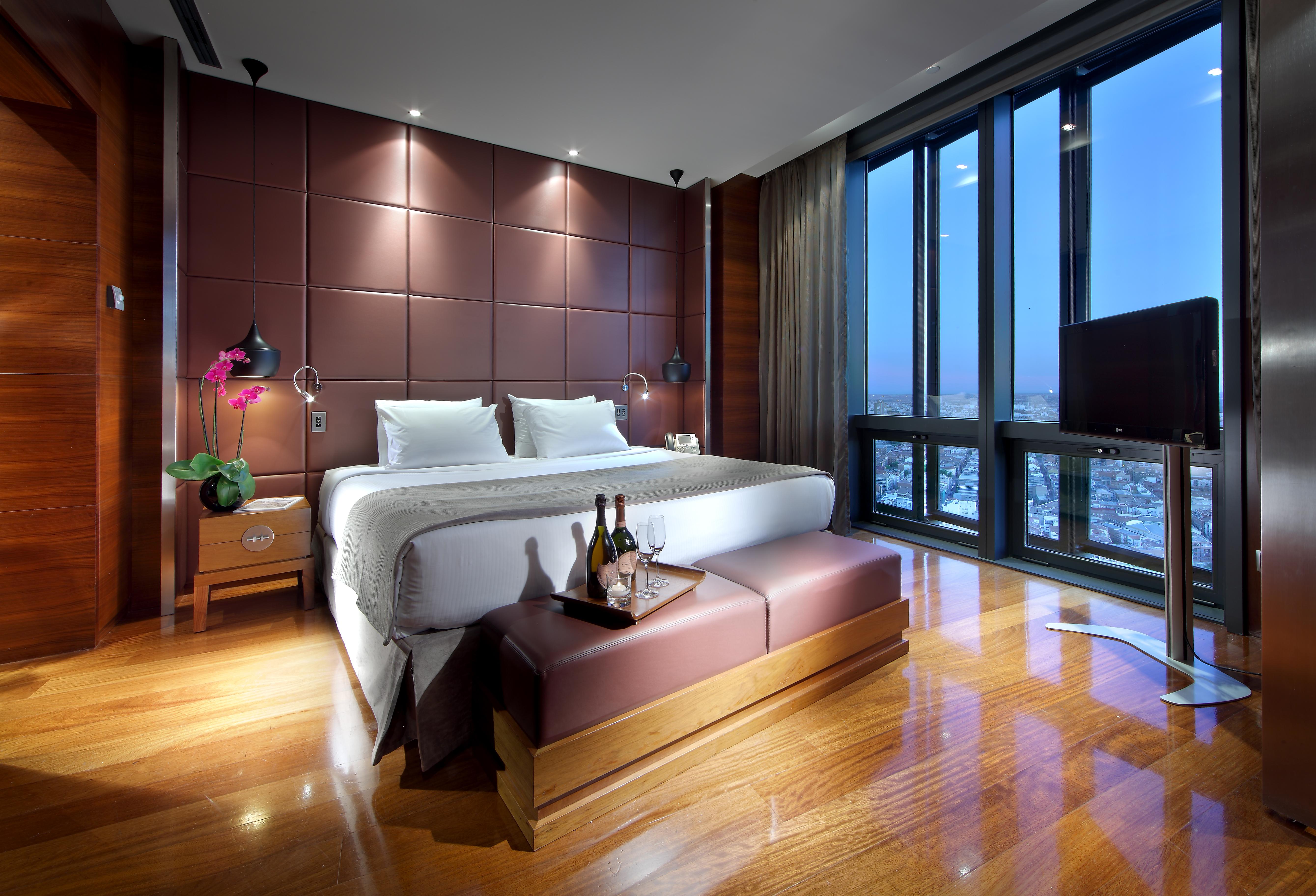 Hotel Eurostars Madrid Tower Esterno foto The photo shows a modern hotel room with a luxurious design. It features a large bed with white linens and plush pillows, positioned against a stylish brown accent wall. There are bedside tables on each side of the bed, each equipped with a lamp. In 