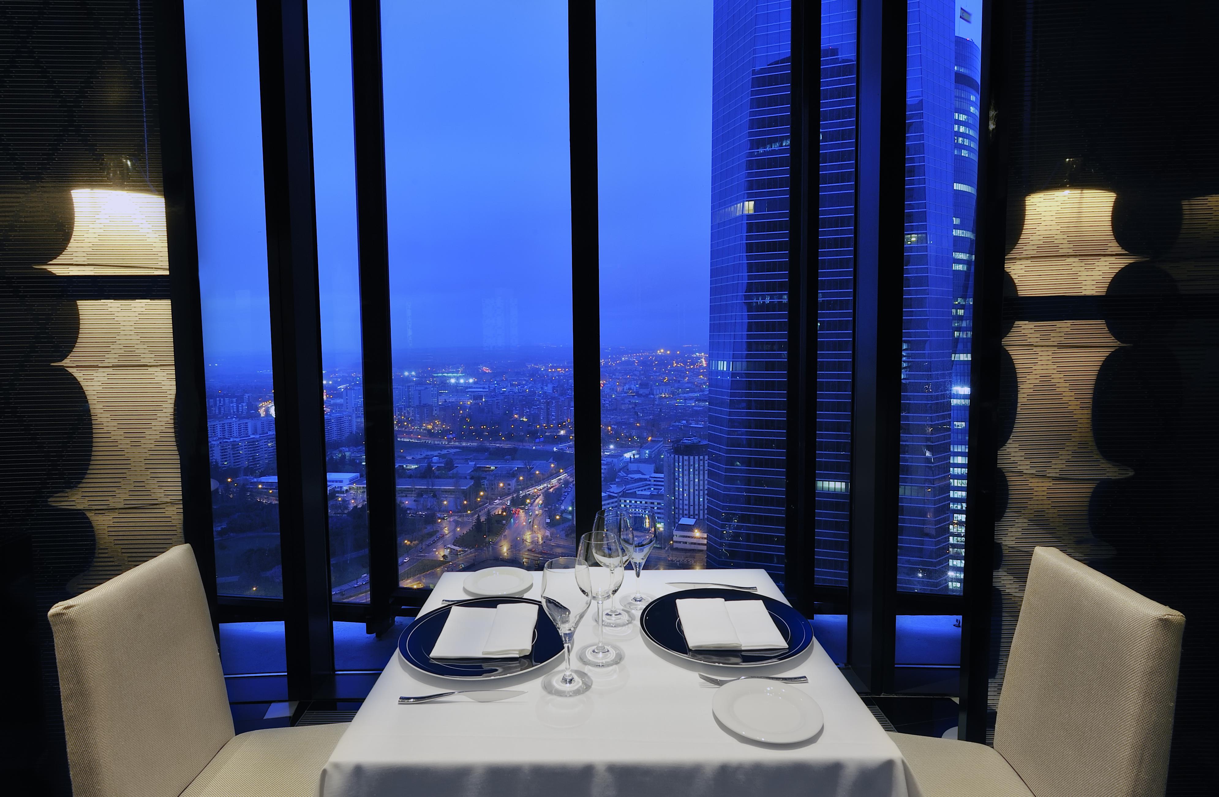 Hotel Eurostars Madrid Tower Ristorante foto The photo shows a fine dining restaurant setting with a table for two, elegantly set with white tablecloth, plates, glasses, and utensils. The table is positioned near large windows that offer a panoramic view of a cityscape at dusk, illuminated by l