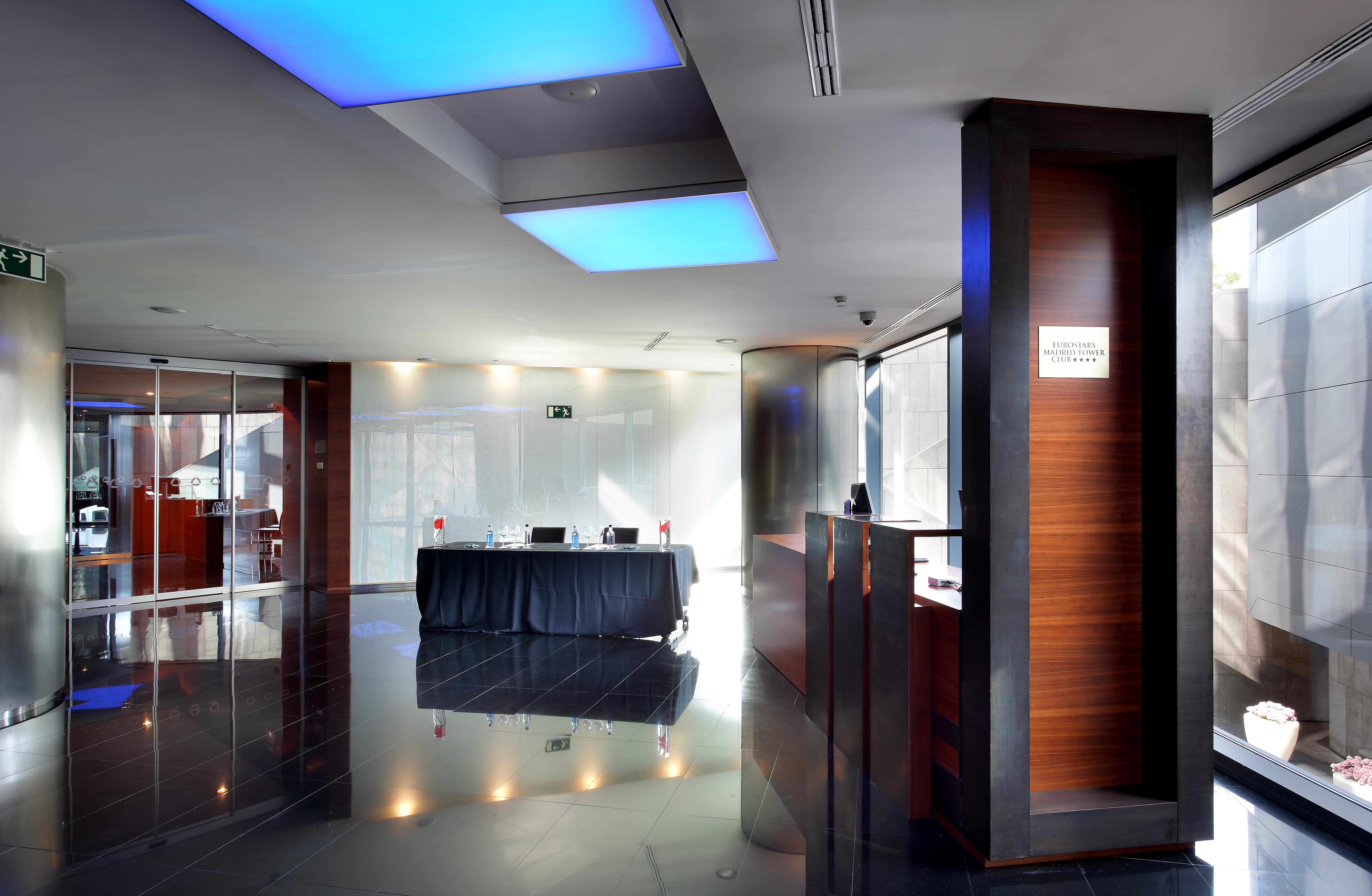 Hotel Eurostars Madrid Tower Esterno foto The image shows the interior of a modern reception area. The space features sleek design elements, including glossy black flooring and walls. There is a front desk made of wood, which includes a sign, likely indicating reception services. Soft blue l