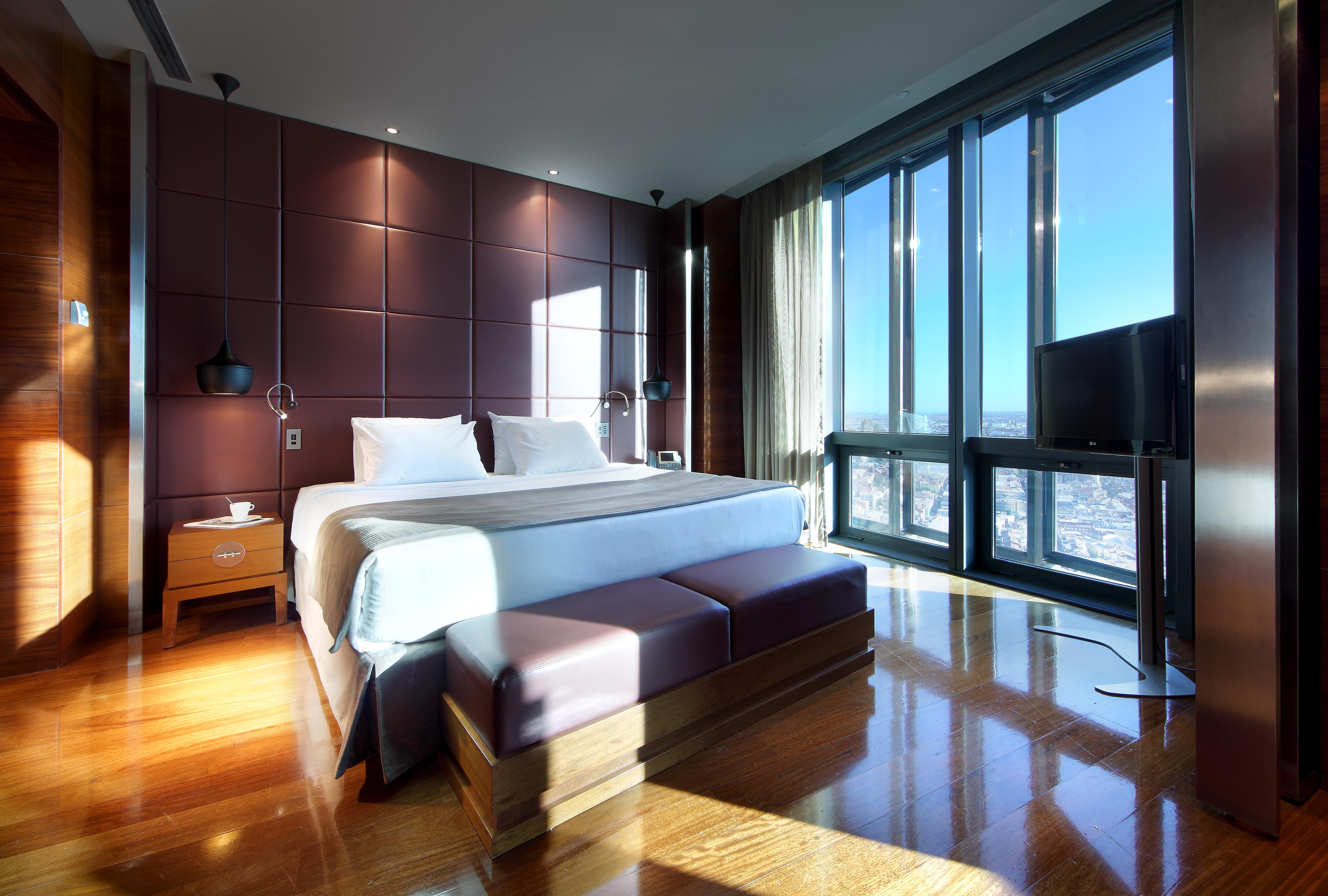 Hotel Eurostars Madrid Tower Esterno foto The photo shows a modern hotel room with sleek design features. It includes a large bed with white bedding, positioned against a wall with dark, textured panels. On either side of the bed, there are bedside tables with lamps. A stylish bench sits at 