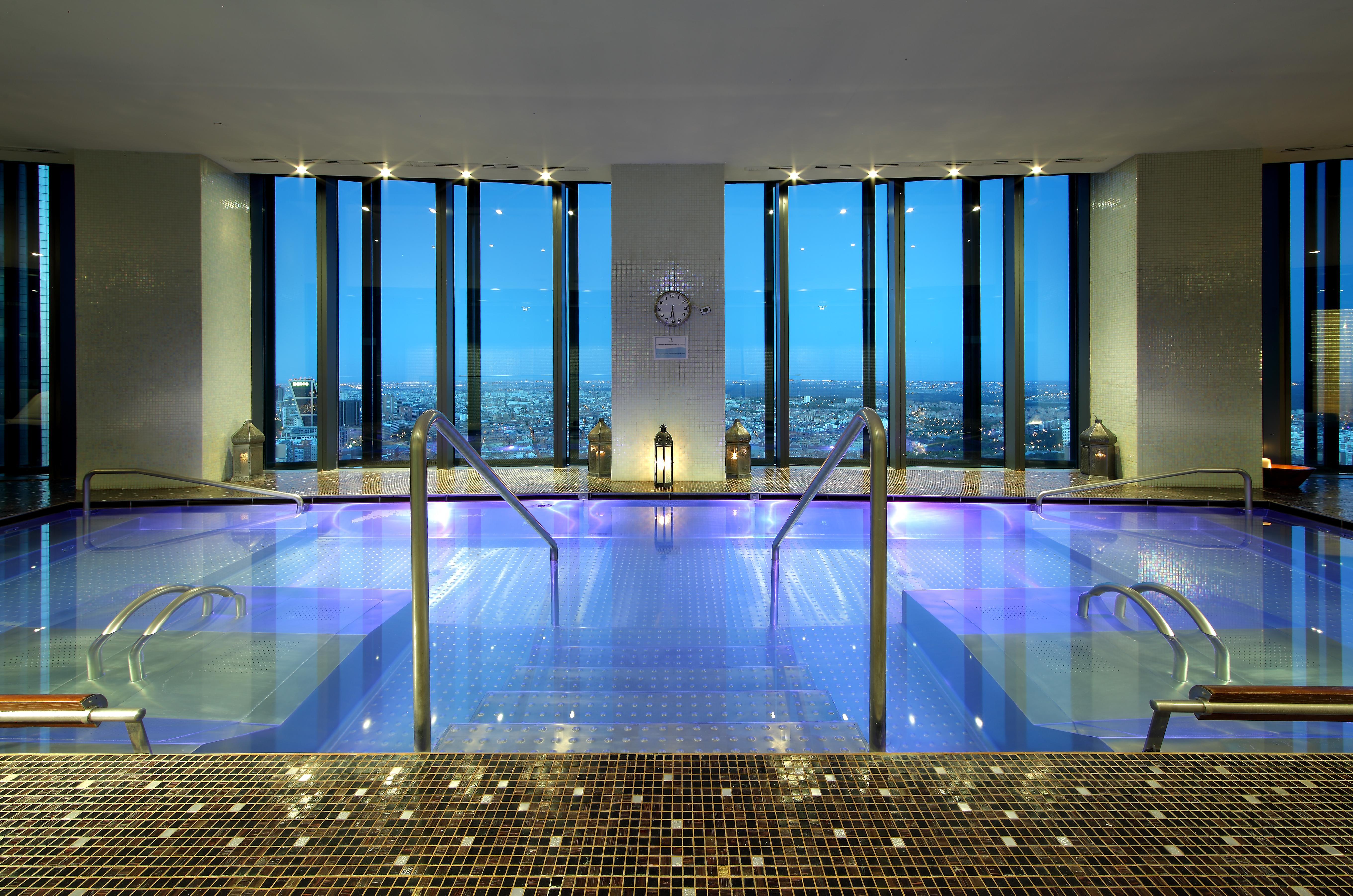 Hotel Eurostars Madrid Tower Esterno foto The image shows a luxurious spa area with a large, tranquil pool that has a vibrant blue hue. The pool is surrounded by sleek, modern design elements, including steps leading down into the water. Large windows provide a stunning view of the cityscape