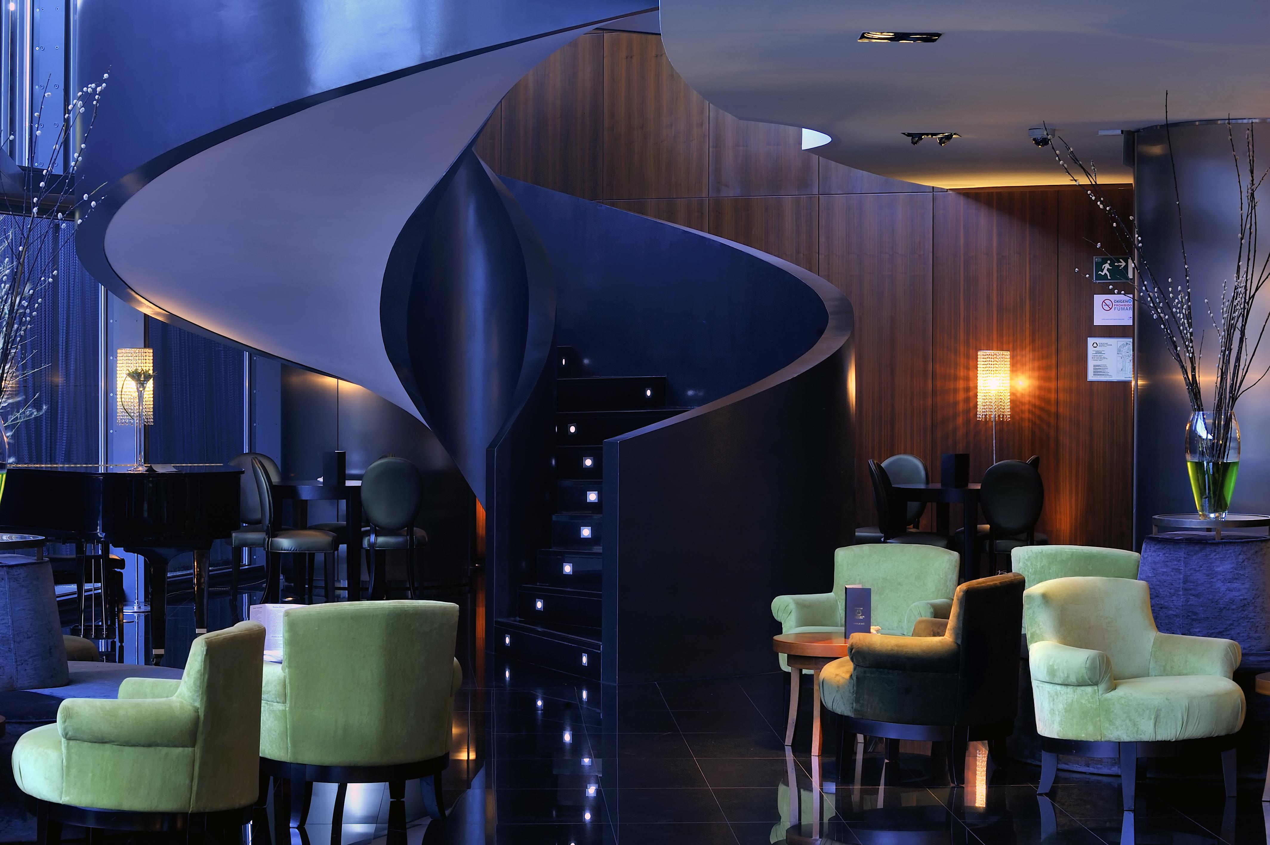 Hotel Eurostars Madrid Tower Esterno foto The photo shows a stylish interior of a modern lounge or hotel lobby. There is a striking spiral staircase that leads to an upper level, illuminated by soft lights along the steps. The color palette features dark tones, including a deep blue wall and