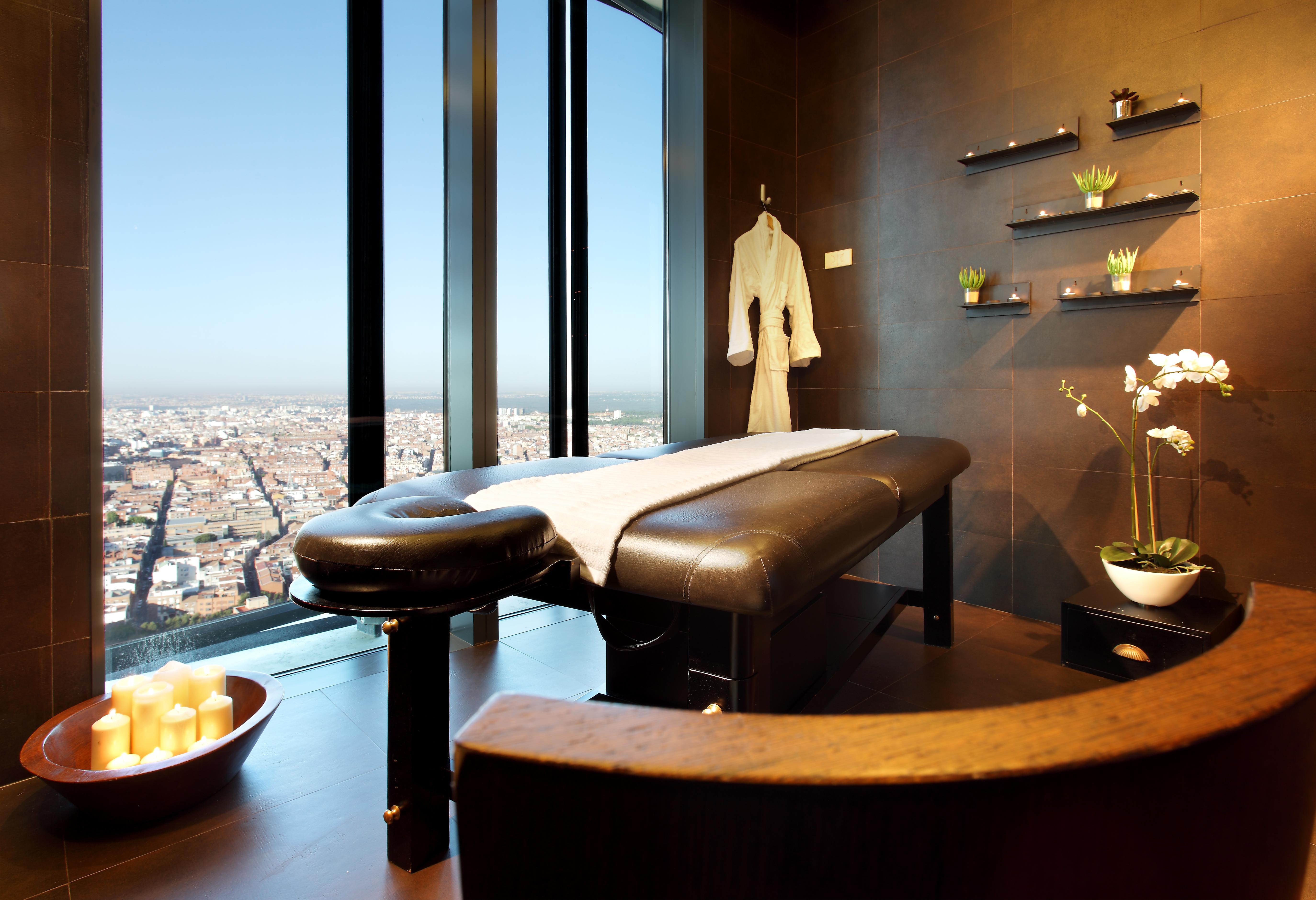 Hotel Eurostars Madrid Tower Esterno foto The photo shows a modern spa treatment room with large windows offering a panoramic view of the city. In the center, there's a massage table covered with a white sheet. A plush bathrobe hangs on the wall, and there are decorative shelves with small p