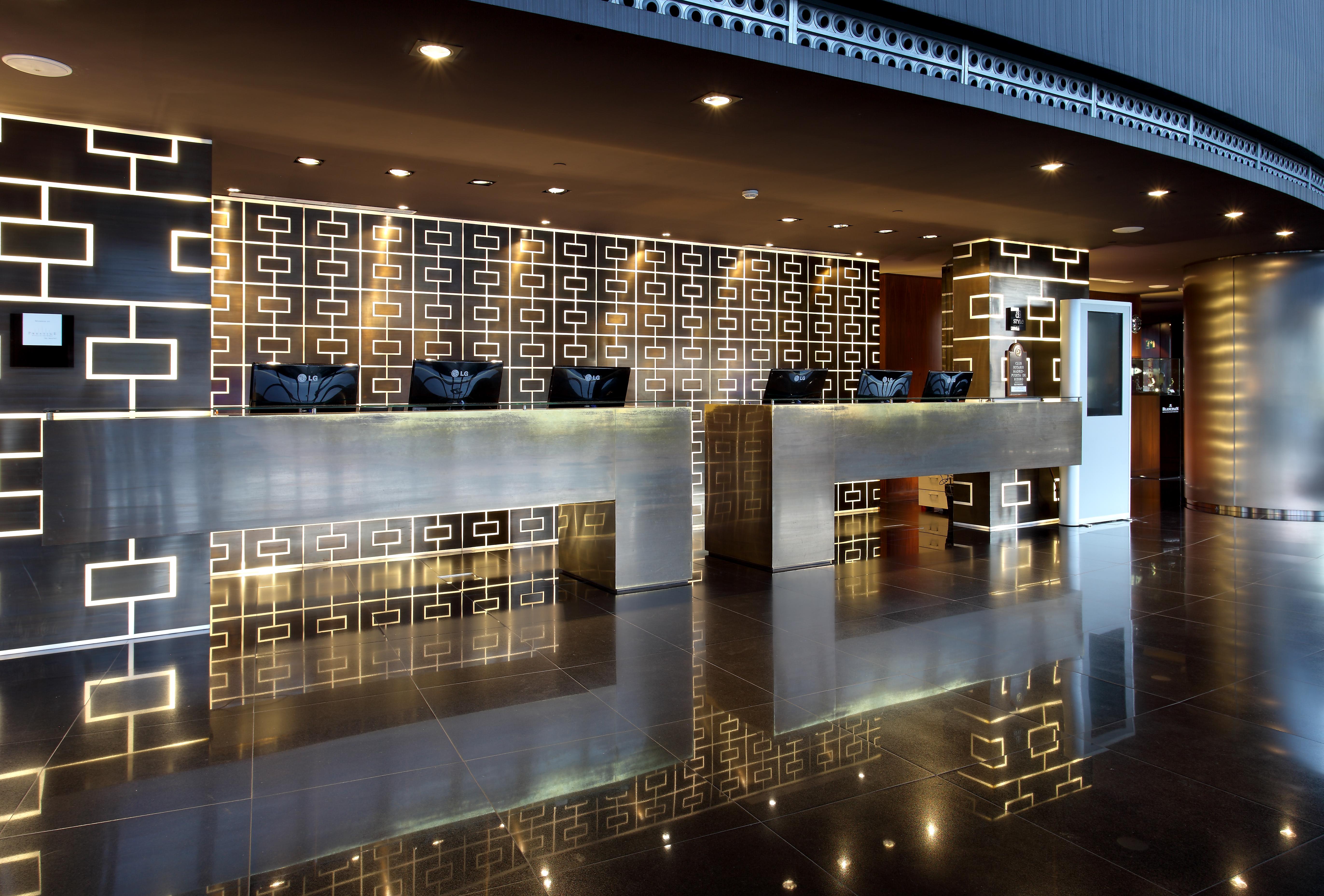 Hotel Eurostars Madrid Tower Esterno foto The photo shows a modern hotel reception area. There are two prominent reception counters made of a sleek, shiny material. The background features a decorative wall with a geometric pattern, possibly in darker colors. The lighting is soft and stylish
