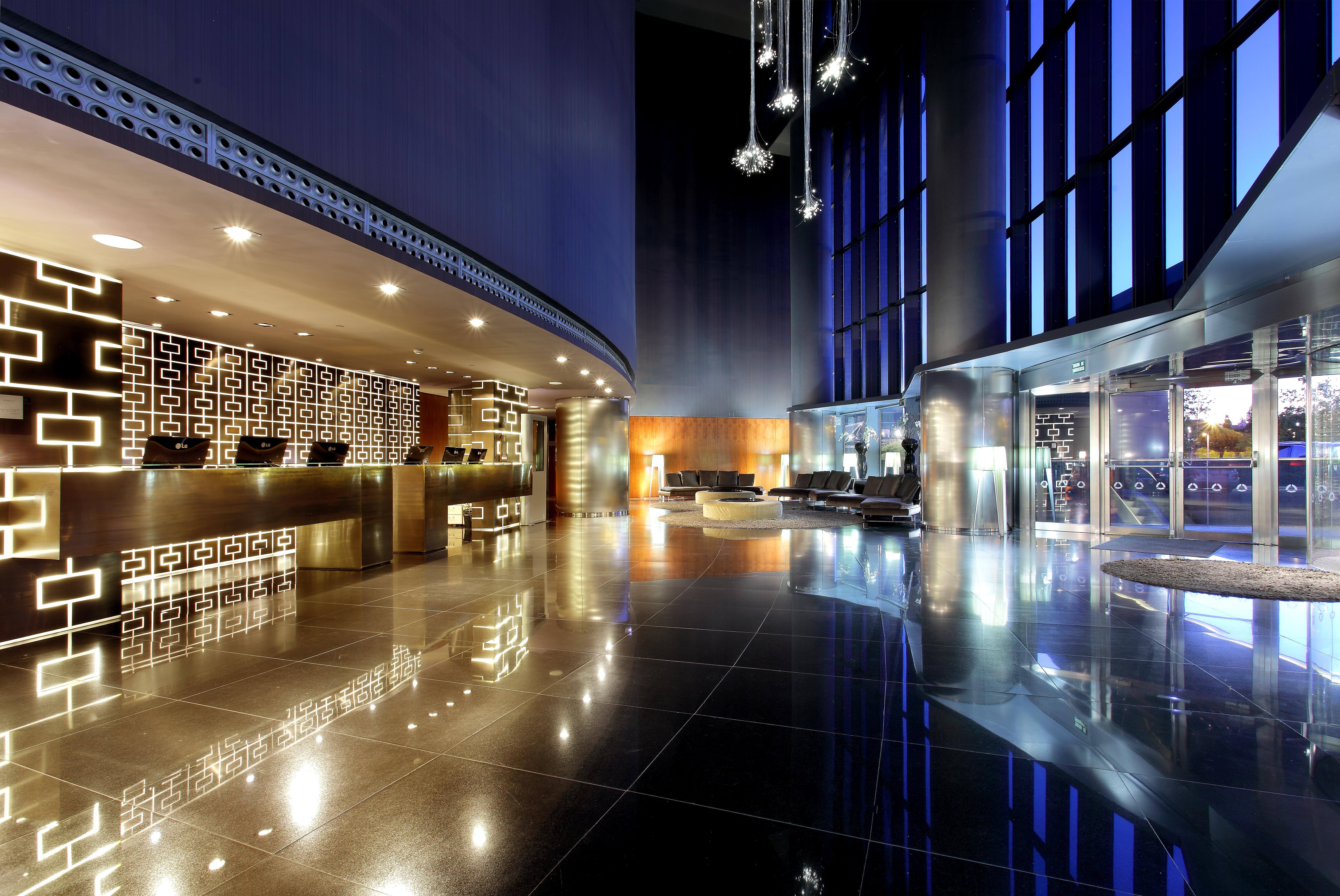 Hotel Eurostars Madrid Tower Esterno foto The image depicts a modern hotel lobby featuring sleek, contemporary design elements. On one side, there is a reception desk with a striking geometric pattern, illuminated with soft lighting. The flooring is glossy and reflective, adding to the elega