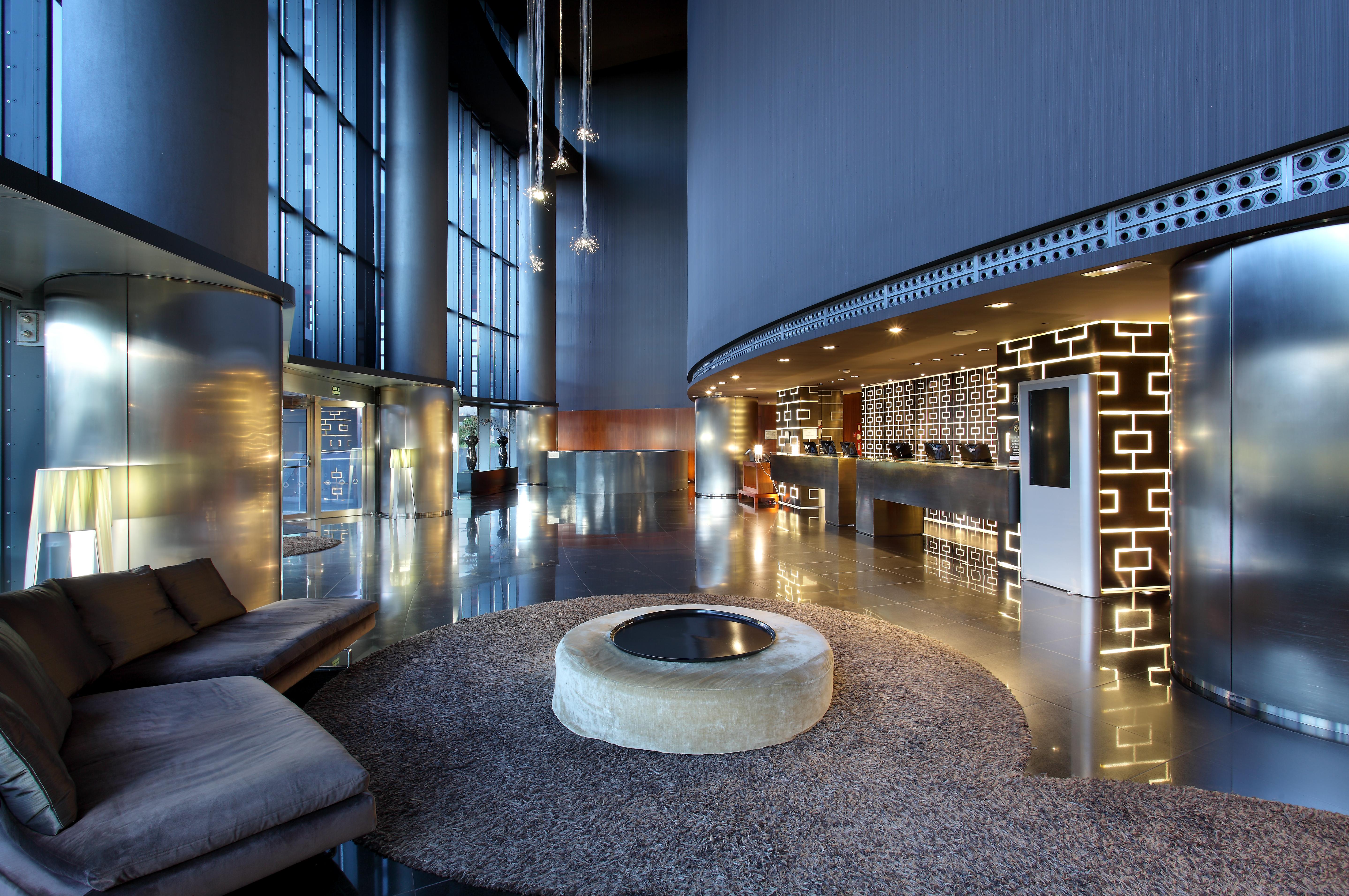 Hotel Eurostars Madrid Tower Esterno foto The photo shows a modern hotel lobby with a sleek and sophisticated design. There is a large, open space featuring tall glass windows allowing natural light to flood in. The reception desk is stylishly designed, with a decorative wall behind it that 