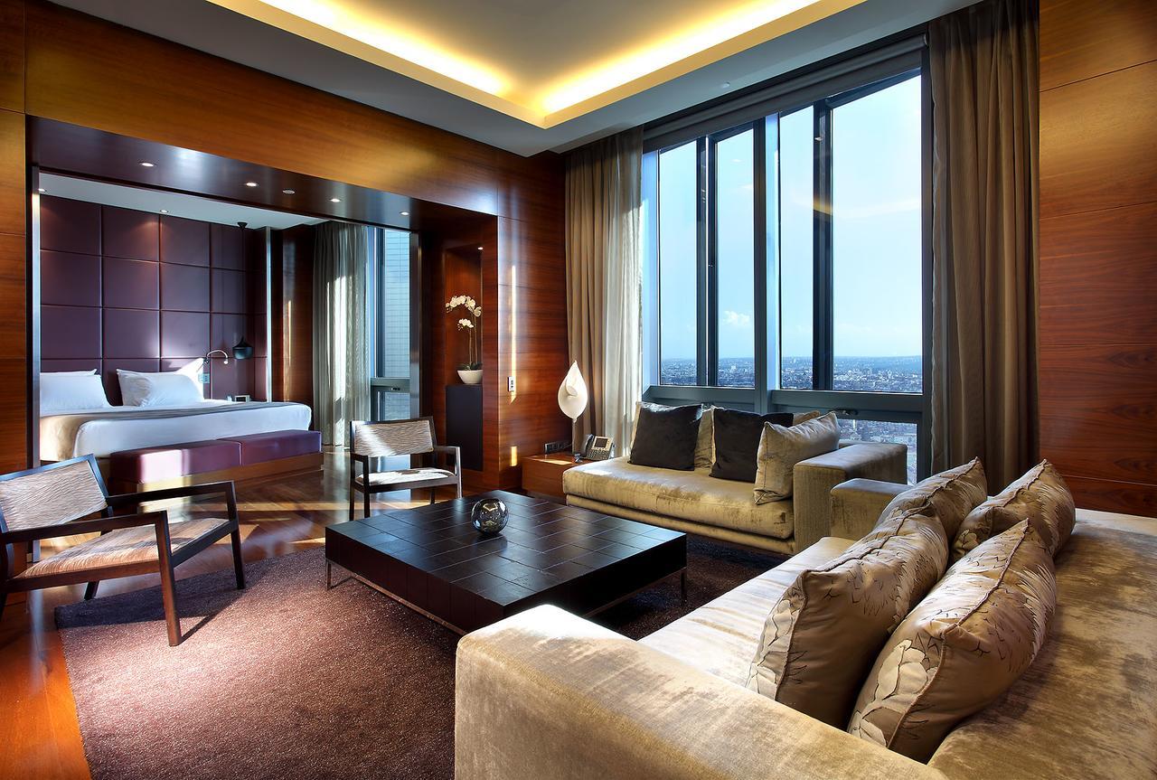 Hotel Eurostars Madrid Tower Esterno foto The photo depicts a modern hotel room featuring a well-furnished living area and a bedroom space. The room has large windows that allow natural light to fill the space, providing a view of the city. The living area includes a plush sofa with decorati