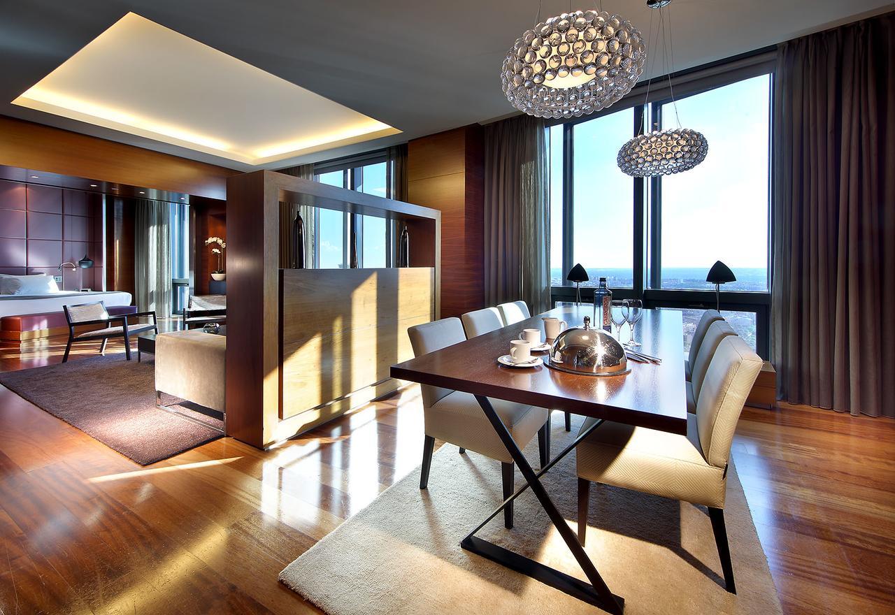 Hotel Eurostars Madrid Tower Esterno foto The photo shows a modern dining area within a spacious interior. A sleek wooden dining table is set for a meal, featuring white dining chairs. Above the table, there are stylish chandelier lights illuminating the space. Large floor-to-ceiling windows