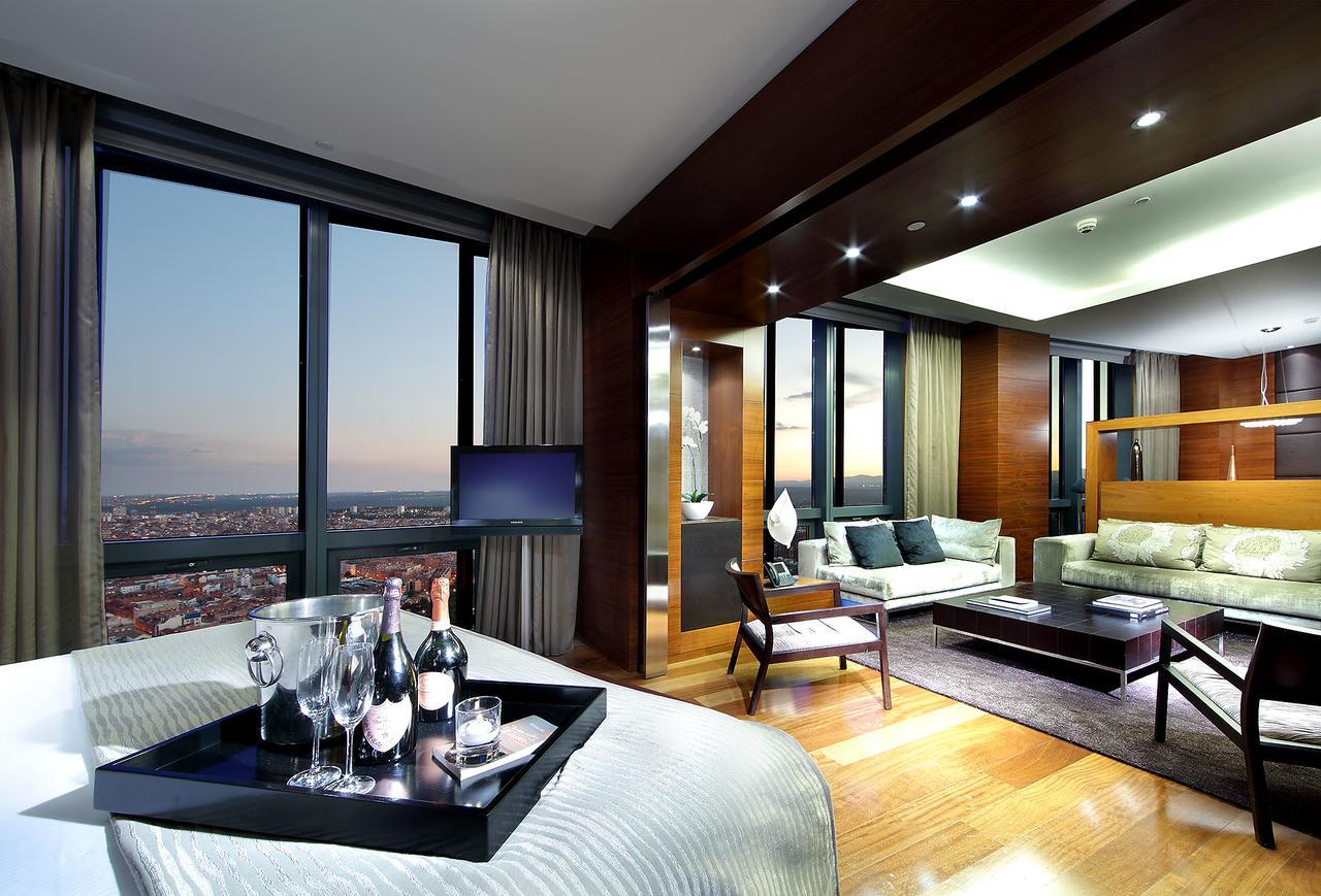 Hotel Eurostars Madrid Tower Esterno foto The photo shows a luxurious hotel suite with modern decor. On one side, there is a bedroom area featuring a large bed, while on the other side, there is a spacious living area with a comfortable sofa and coffee table. The room is well-lit with stylis