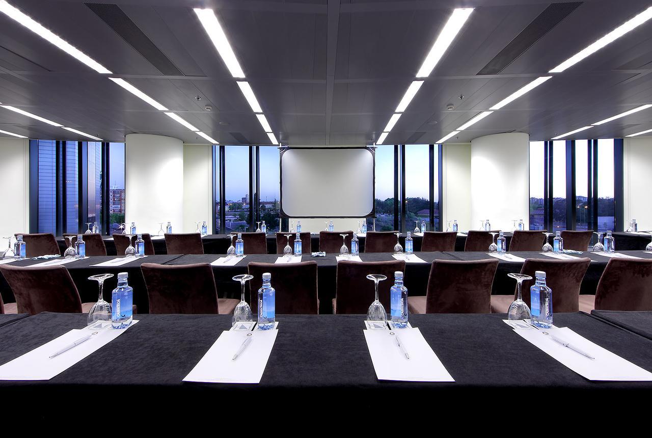 Hotel Eurostars Madrid Tower Esterno foto The photo shows a modern conference room with large windows that provide a view of the cityscape outside. The room is arranged with several tables set up in a classroom or boardroom style. Each table has a black tablecloth and features a glass of wat