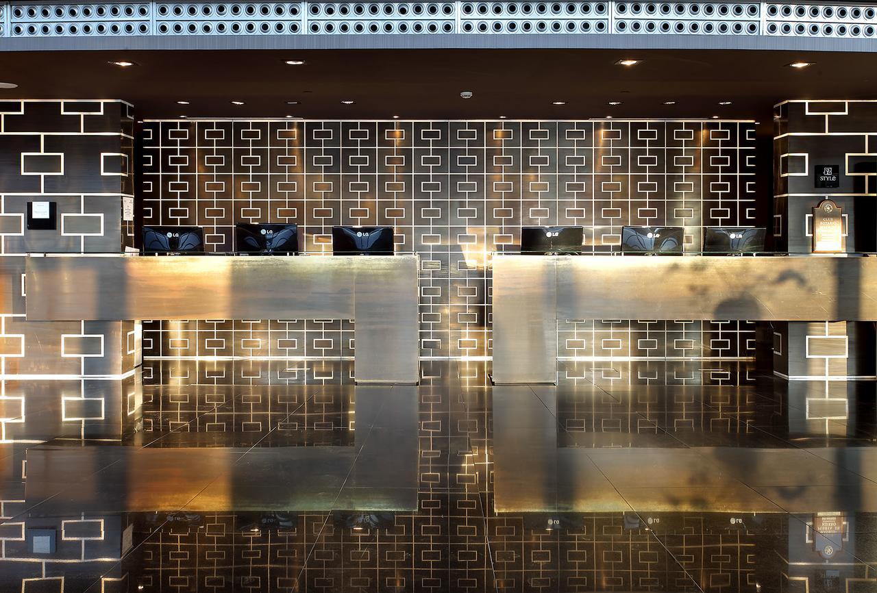 Hotel Eurostars Madrid Tower Esterno foto The photo shows a modern reception area or lobby. It features two sleek reception desks made of a polished, metallic material that reflects light. Behind the desks is a decorative wall with a geometric pattern in dark tones. The overall design is con