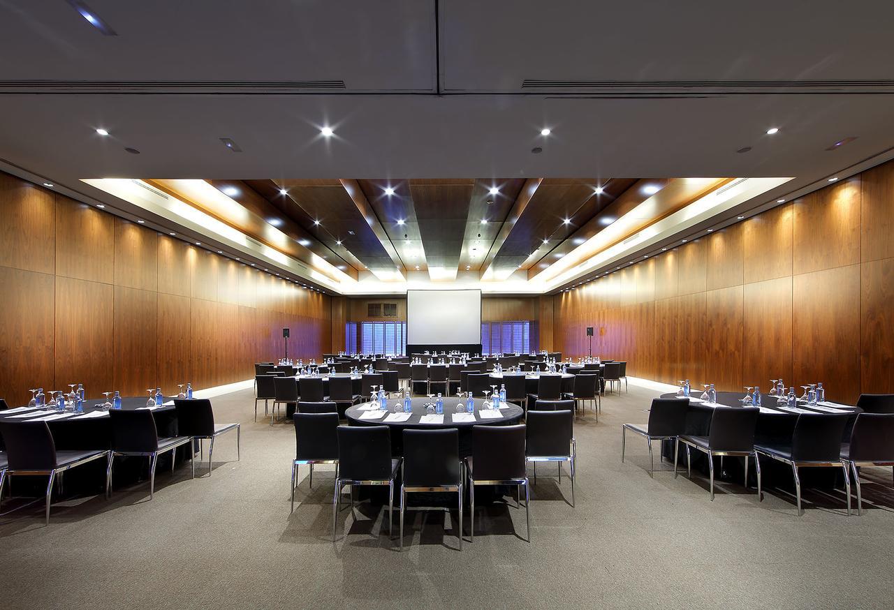 Hotel Eurostars Madrid Tower Esterno foto The photo shows a spacious and modern conference room set up for a meeting or event. There are several round tables arranged in a semi-circle, each covered with a white tablecloth and equipped with water glasses, writing pads, and pens. The room feat