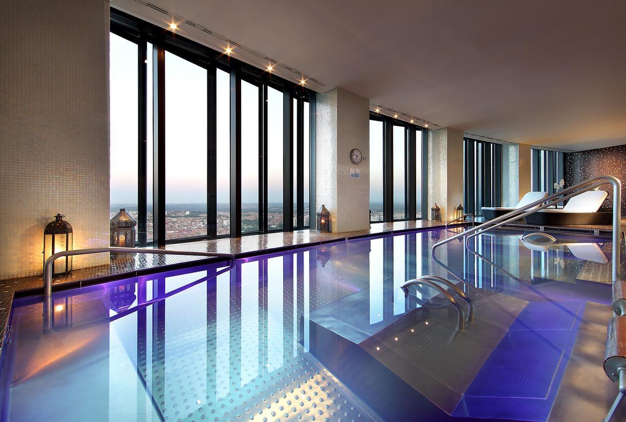 Hotel Eurostars Madrid Tower Esterno foto The photo shows a luxurious indoor pool area with large floor-to-ceiling windows that provide a stunning view of the skyline. The pool features a shiny, reflective surface and has a modern design, with gentle curves in the structure. The lighting is 