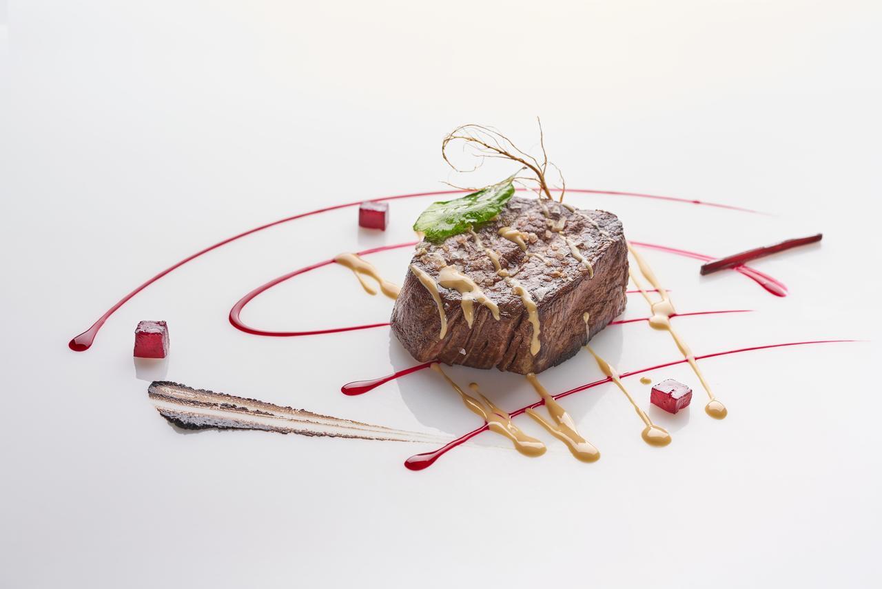 Hotel Eurostars Madrid Tower Esterno foto The photo features a beautifully plated dish that showcases a piece of meat, likely a steak, at the center. The meat is glazed with a sauce and garnished with a thin piece of green herb. Surrounding the meat are artistic drizzles of different sauces,