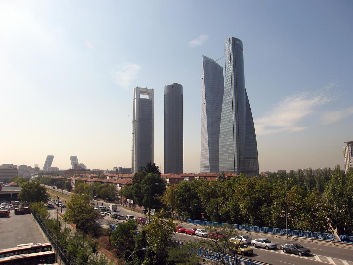 Hotel Eurostars Madrid Tower Esterno foto The photo shows a cityscape featuring several modern skyscrapers, prominently four tall buildings in the foreground. The structures have distinctive architectural designs, with varying shapes and heights. The scene includes a tree-lined area and a bu