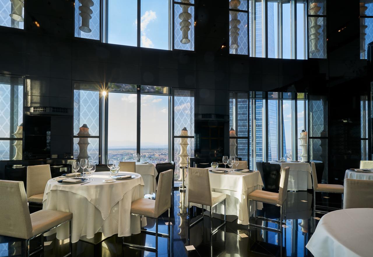 Hotel Eurostars Madrid Tower Esterno foto The photo features an elegant restaurant interior with large windows that provide a panoramic view of the city skyline. The tables are elegantly set with white tablecloths and fine dining tableware, including glasses and plates. The decor appears mod