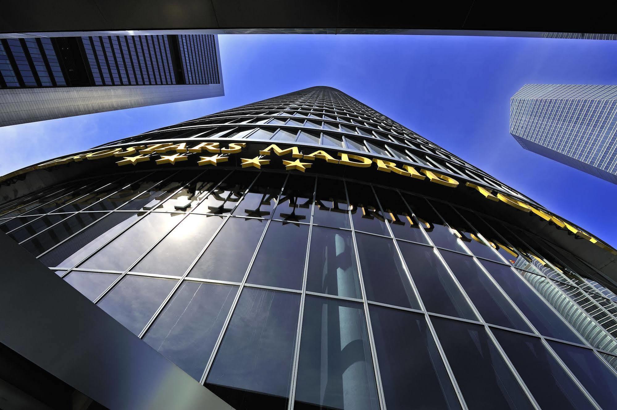 Hotel Eurostars Madrid Tower Esterno foto The photo shows a tall modern building reaching towards the sky. The perspective is from the ground looking upward, highlighting the building's height and glass facade. The building features a large sign that reads "MADRID" in bright yellow letters, 