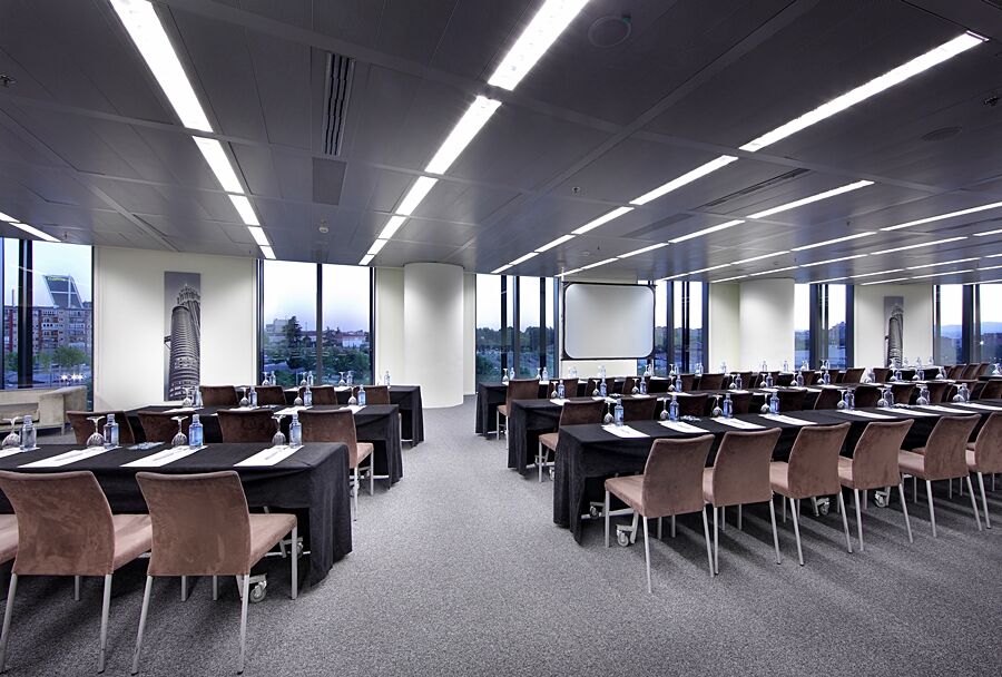 Hotel Eurostars Madrid Tower Esterno foto The photo shows a modern conference room set up for a meeting or seminar. There are multiple rows of tables arranged in a classroom style, each covered with black tablecloths. On the tables, there are notepads and water bottles, suggesting that atten